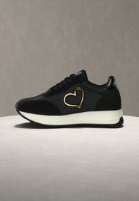 Μαύρο αθλητικό παπούτσι με λευκή σόλα, μαύρα κορδόνια και χρυσή καρδιά σε σχήμα διακοσμητικό στο πλάι, τοποθετημένο σε ουδέτερη επιφάνεια.