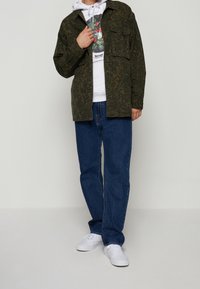 Olivgrön mönstrad skjortjacka över en vit grafisk hoodie, i kombination med blå jeans och vita sneakers. Enkel rund halsringning och två bröstfickor.