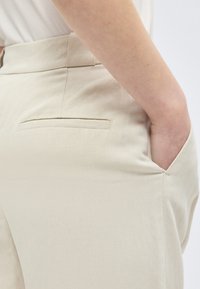 Inimese lähivõte, kes kannab beeže pükse, ühe käega ees taskus, kuvatakse külgede ja tagataskute detailid.