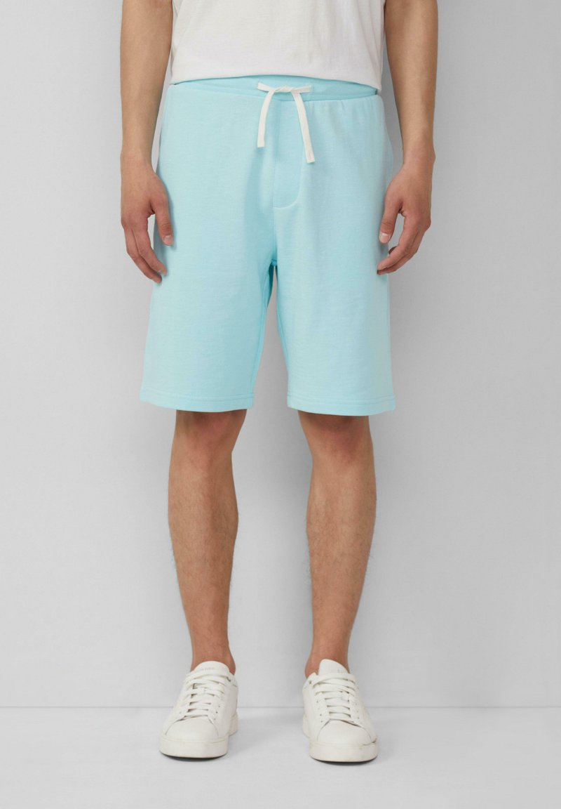 Helle blaue Baumwollshorts mit einem Kordelzugbund, seitlichen Taschen und umgenähten Kanten, kombiniert mit weißen Turnschuhen auf einem neutralen Hintergrund.