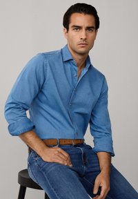 Lichtblauwe denimshirt met knoopsluiting, lange mouwen, opgerolde manchet en een subtiele merkl logo op de borst. Draagt met donkere jeans en een bruine riem.