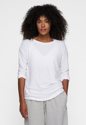 Camisa blanca de manga larga hecha de una tela suave con un escote amplio y mangas arremangadas, combinada con pantalones deportivos gris claro que tienen una etiqueta de acento.