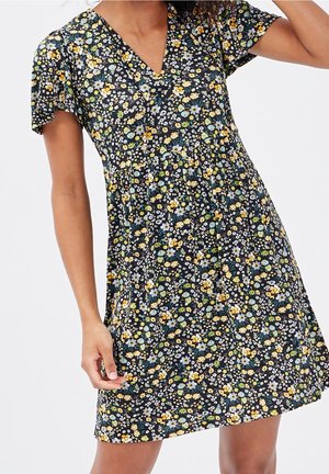 Femme portant une robe à manches courtes à fleurs bleues, jaunes, blanches et vertes sur fond noir.