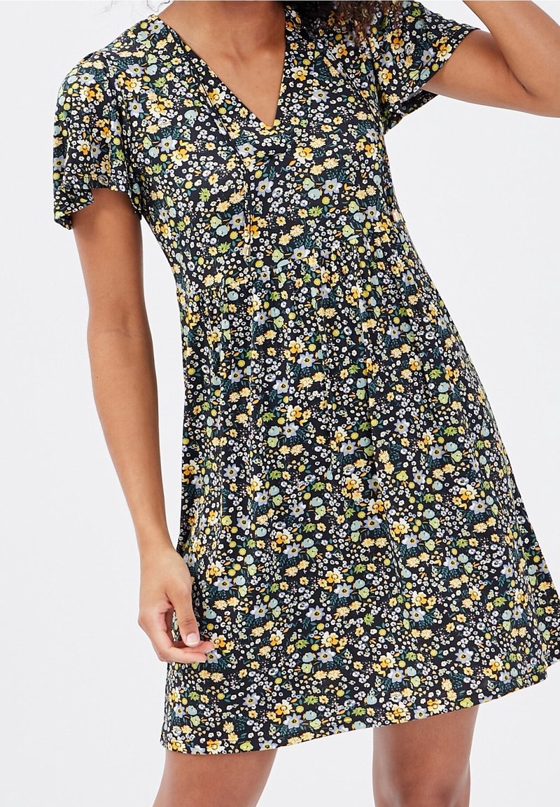 Femme portant une robe à manches courtes à fleurs bleues, jaunes, blanches et vertes sur fond noir.