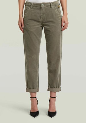 Olivgrüne Cordhosen, an den Knöcheln aufgerollt, getragen mit schwarzen Pumps mit Knöchelriemen und spitz zulaufender Schuhspitze, gezeigt von der Taille bis zu den Füßen.