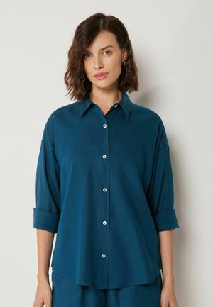 Kobieta o brązowych włosach długości do ramion, ubrana w luźną zielonkawo-niebieską koszulę na guziki z podwiniętymi rękawami, na tle jasnego, gładkiego tła.