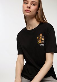 Czarny bawełniany t-shirt z kwiatowym wzorem w odcieniach żółtego i pomarańczowego po lewej stronie klatki piersiowej, z napisem "apoh London" wydrukowanym poniżej. Krótkie rękawy.