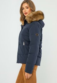 Granatowa kurtka puchowa z kapturem wykończonym sztucznym futrem, zapinanymi kieszeniami i teksturowanym materiałem, z naszywką logo na rękawie.