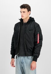 Młody mężczyzna w czarnej kurtce bomber z czerwoną metką "Remove Before Flight" i szarym dzianinowym swetrze, stojący na białym tle.