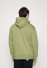 Felpa color verde oliva con tasca a marsupio, cappuccio regolabile con lacci e polsini a coste. Presenta un logo sulla manica sinistra.