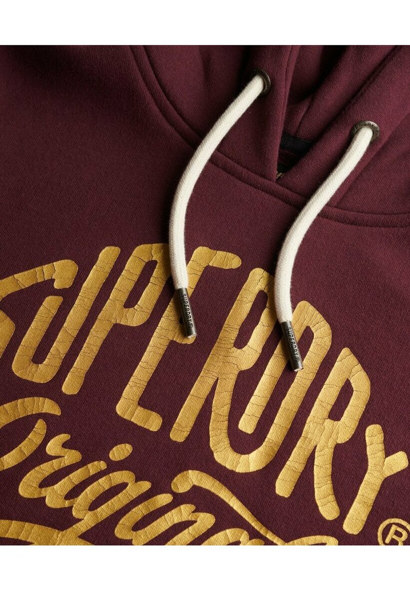 Bordowa bluza z kapturem z teksturowanym złotym nadrukiem z napisem "SUPERDRY" oraz "Originals". Białe sznurki z metalowymi końcówkami dopełniają całość.