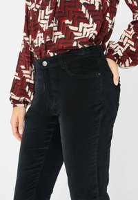 Zwarte corduroy broek met een gladde textuur, voorzien van steekzakken en een knoopsluiting. G gedragen met een gebloemde blouse.