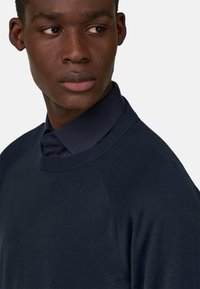 Jeune homme noir portant un pull bleu marine par-dessus une chemise à col bleu foncé, regardant vers sa gauche sur un fond clair uni.