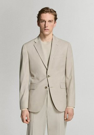 Hombre joven de pie, vestido con una chaqueta de traje beige y pantalones a juego, con una camiseta beige de cuello redondo debajo, frente a un fondo claro y liso.