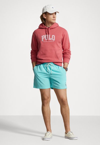 Piros kapucnis pulóver "Polo Ralph Lauren" logóval, világoskék fürdőnadrággal párosítva. Kiegészítve egy fehér sapkával és bézs sportcipőkkel.