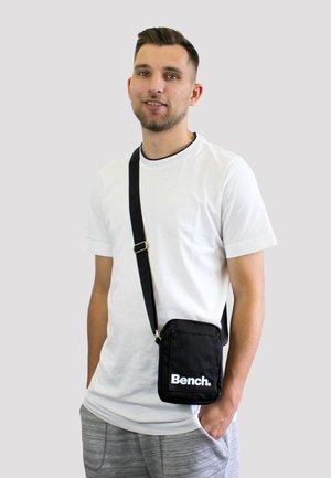 Schwarze Stoff-Umhängetasche mit einer vorderen Reißverschlusstasche. Das Wort "Bench." ist in Weiß auf der Vorderseite aufgedruckt. Verstellbarer Tragegurt und rechteckige Form.