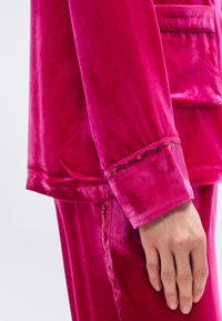 Χέρι που ακουμπά δίπλα στο ισχίο, φορώντας φωτεινό ροζ σατέν σακάκι και παντελόνι με παγιέτες στα μανίκια και την άκρη της τσέπης.