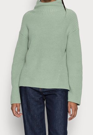 Un pull en tricot vert menthe avec un col haut et des épaules tombantes. Présente une texture côtelée et des manches longues, assorti à un jean sombre.