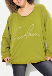Sweatshirt oversized vert olive avec des poignets côtelés et un décolleté large, arborant un texte brodé blanc "Freedom" et "Est. 1981" sur le devant.