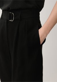 Pantalons noirs en tissu texturé, taille ceinturée, poches latérales et coupe ajustée. Les détails de surpiqûres visibles rehaussent le design.