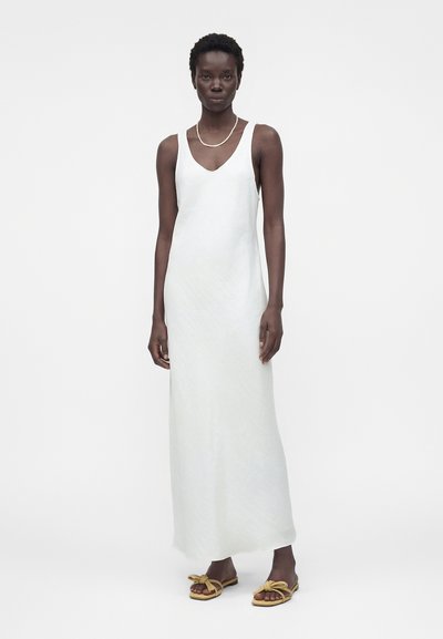 Γυναίκα που φοράει λευκό maxi φόρεμα χωρίς μανίκια, χρυσό κολιέ και μπεζ σανδάλια με δεμένους ιμάντες, στέκεται μπροστά σε μια απλή ανοιχτόχρωμη επιφάνεια.