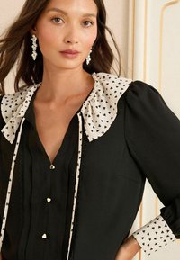 Donna che indossa una blusa nera con colletto e polsini arricciati a motivo a cuore bianco, bottoni a forma di cuore e orecchini pendenti in cristallo.