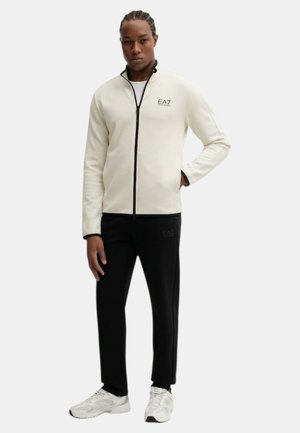 Uomo con capelli intrecciati che indossa una giacca bianca con cerniera e pantaloni neri con scarpe da ginnastica bianche, in piedi con una mano in tasca.