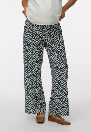 Femme portant un pantalon à jambes larges avec un motif de feuilles bleu et blanc, associé à des chaussures plates marron et un haut blanc.