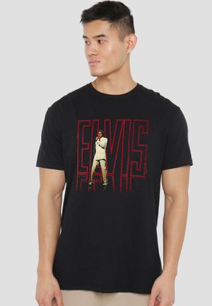 Junger Mann trägt ein schwarzes T-Shirt mit rotem "ELVIS"-Schriftzug und einem Bild von Elvis Presley im weißen Anzug singend auf der Vorderseite.