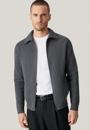 Mężczyzna w ciemnoszarej kurtce na zamek, białej koszulce i czarnych spodniach z paskiem, stoi na jednolitym jasnym tle.