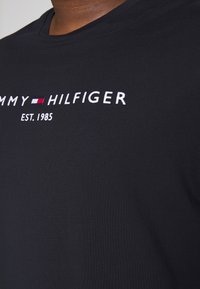 Černé bavlněné tričko s výšivkou loga "TOMMY HILFIGER" a "EST. 1985" v bílé barvě s malými červenými a modrými akcenty. Krátké rukávy.