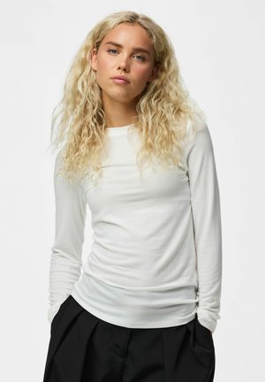 Jeune femme aux longs cheveux blonds et bouclés portant un haut blanc moulant à manches longues et un pantalon noir ample, debout les mains dans les poches, expression neutre.