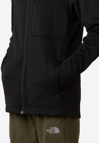 Black fleece jacket featuring a textured fabric, zippered front, side pockets, and an embroidered logo on the lower corner.