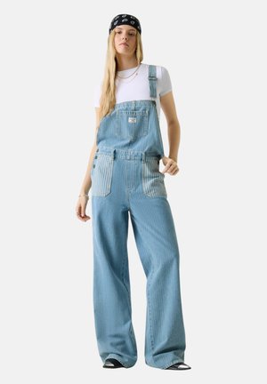 Giovane donna che indossa una salopette di jeans azzurro chiaro con tasche a righe, una maglietta bianca a maniche corte, una bandana nera e gioielli d'argento, in piedi dritta.