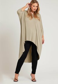 Khaki oversized topp med vida ärmar, böjd fåll och sidslitsar, kombinerad med svarta tajta byxor och svarta sandaler med remmar.