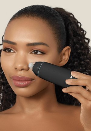 Kvinde med krøllet hår bruger en sort ansigtsrens til porer på sin højre kind mod en neutral baggrund.