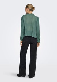 Γυναίκα με ξανθό κότσο που φοράει διάφανη πράσινη μπλούζα με μακριά μανίκια και μαύρο τζιν με φαρδιά παντελόνια, στέκεται με την πλάτη προς τα εμπρός σε απλό φόντο.