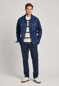 Džinsinė striukė su mygtukais, dviem priekinėmis kišenėmis ir mėlyna spalva; derinama su kremine megztine su mėlynomis juostelėmis ir tamsiomis kelnių.