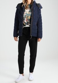 Marinblå puffjacka med dragkedja fram, grå krage, blommig tröja, svarta smala byxor och vita sneakers. Slätt material och quiltat mönster.