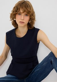 Top senza maniche blu navy con scollatura strutturata e arrotondata, con dettagli plissettati, abbinato a jeans in denim. Tessuto liscio con design aderente.