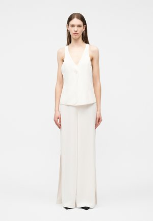 Žena stojaca, oblečená v bielej bezrukávovej blúzke s výstrihom do V a jedným gombíkom a v zodpovedajúcich širokých nohaviciach na jednoduchom pozadí.