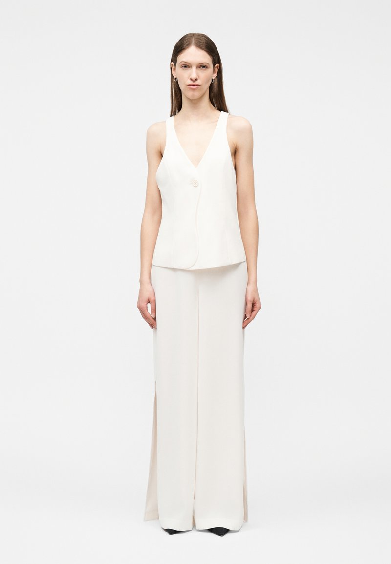 Γυναίκα όρθια, φοράει λευκό αμάνικο τοπ με V-λαιμόκοψη και ένα κουμπί, καθώς και ταιριαστό παντελόνι με φαρδιά μπατζάκια, μπροστά σε απλό φόντο.