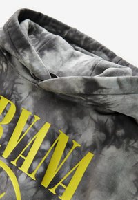 Šedá mikina s kapucí v tie-dye stylu, zdobená velkým žlutým nápisem "Nirvana", měkkou texturou a uvolněným střihem s zavinutou kapucí.