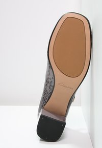 Černá texturová podrážka boty s vzorem hadí kůže, s hnědou gumovou základnou a průhledným podpatkem. Na straně je vyražené logo "Clarks".