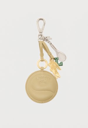 Porte-clés rond en cuir beige avec logo Lacoste, breloque crocodile dorée, breloque "L" verte et breloque raquette de tennis argentée sur un clip métallique.