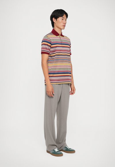 Polo a righe in tonalità multicolori con colletto bordeaux, abbinato a pantaloni grigi chiari plissettati e sneakers verdi con lacci bianchi.
