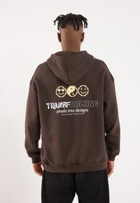 Brown hoodie with a front kangaroo pocket, featuring pixelated graphics and white text on the back. Soft fabric, relaxed fit.
