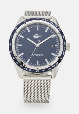 Ceas Lacoste cu un cadran rotund de culoare navy, brățară din plasă argintie, indicatoare de oră argintii și bezel tachimetru. Dispune de o fereastră pentru dată la ora 3.