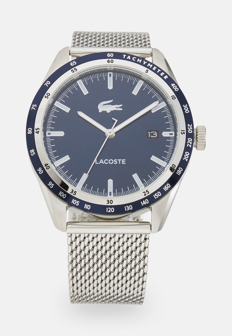 Lacoste watch with a round navy dial, silver mesh band, silver hour markers, and tachymeter bezel. Features a date window at 3 o'clock.