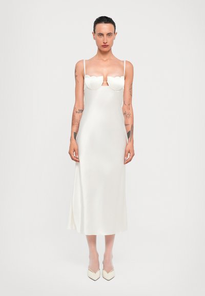 Witte zijden midi-jurk met gegolfd decolleté, dunne bandjes en een zijsplits. Model draagt crème puntige schoenen, met zichtbare lichaamstattoos.